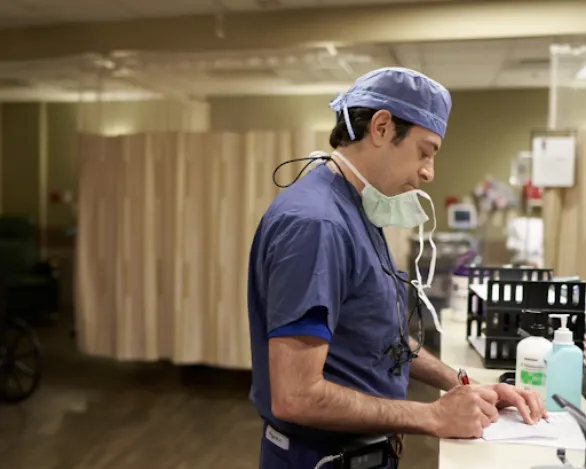 A doctor wearing blue scrubs stands at a desk and writes on a pad of paper