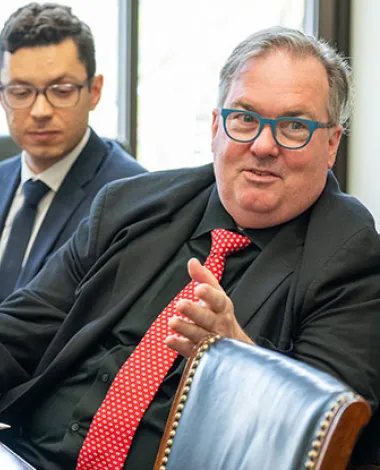Three men sit at a desk talking, with an American flag visible in the background