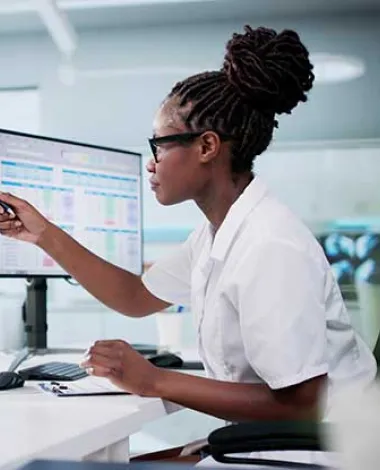 A woman sits at a desk with many monitors looking at charts and spreadsheets