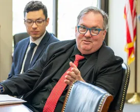 Three men sit at a desk talking, with an American flag visible in the background