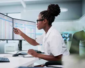 A woman sits at a desk with many monitors looking at charts and spreadsheets