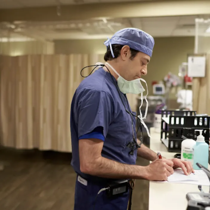 A doctor wearing blue scrubs stands at a desk and writes on a pad of paper