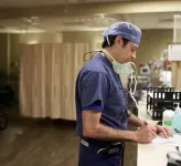 A doctor wearing blue scrubs stands at a desk and writes on a pad of paper
